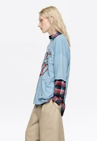 Camiseta de gran tamaño azul claro con diseño gráfico rojo, sobre una camisa a cuadros roja y azul; pantalones cortos de pierna ancha color marrón.