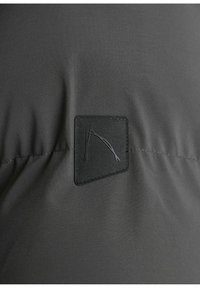 Šedá vonkajšia látka má textúrovaný povrch s štvrtou čiernou nášivkou, na ktorej je minimalizované logo, vyšívané detaily a jemný lesk.