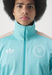 Turkisblå lynlåsjakke med ribbet krave, orange stribede detaljer og hvidt Adidas-logo. Har et rundt Miami klubmærke på brystet.