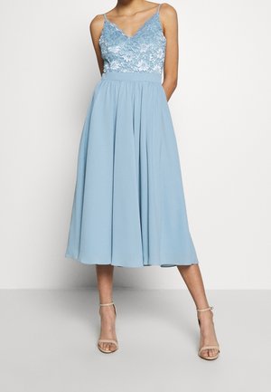 Kobieta ubrana w jasnoniebieską sukienkę midi bez rękawów z koronkowym gorsetem i dopasowaną talią, zestawioną z beżowymi sandałami na wysokim obcasie z paskiem wokół kostki.