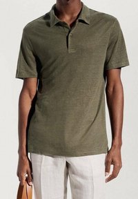 Mann in olivgrünem kurzärmligem Poloshirt und hellbeigen Hosen, hält in einer Hand einen braunen Lederriemen.