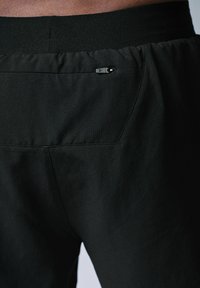 Czarne spodnie sportowe z teksturowanej tkaniny, elastycznym paskiem w talii i małą zapinaną na zamek kieszonką z tyłu. Dopasowany krój i prosty design.