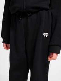 Sorte joggingbukser med elastisk talje og justerbar snor, med en lille hvid bie-logo på venstre lår, båret sammen med en matchende sort hættetrøje.