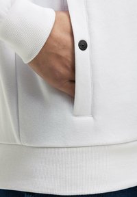 Sudadera blanca con puños y dobladillo acanalados, que presenta un bolsillo lateral y un detalle de botón negro. Textura suave y diseño casual.