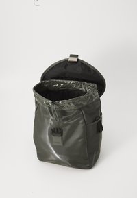 Sac à dos imperméable vert foncé avec un haut à cordon, doté d'un rabat texturé noir, d'un patch logo et d'une sangle réglable. Fini lisse.