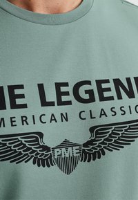 Light green cotton T-shirt featuring bold black text reading "THE LEGEND" and "AMERICAN CLASSIC" with a winged shield graphic.