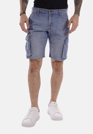 Uomo che indossa pantaloncini cargo in denim azzurro chiaro con tasche a patta e sneakers bianche, in piedi con le mani vicino ai fianchi su sfondo bianco.