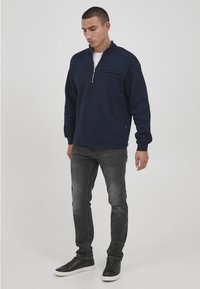 Marineblauer Pullover mit einem Viertel-Reißverschluss-Kragen, der über eine kleine Fronttasche verfügt und aus weichem Material gefertigt ist. Kombiniert mit grauen Jeans und schwarzen Sneakern.
