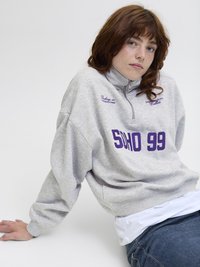 Šedá mikina se zipovým límcem, zdobená fialovým nápisem "SOHO 99" a výšivkovými detaily. Nositá přes bílou košili a v kombinaci s modrými džíny.