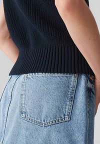 Närbild av en person som bär ljusblå denimjeans och en svart texturerad ärmlös topp, med fokus på bakfickan och midjeområdet.