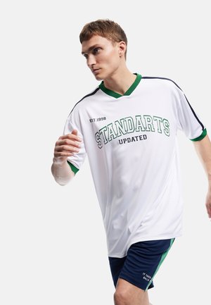 Ung mand i hvidt og grønt sportstøj løber, iført en trøje med teksten "STANDARTS UPDATED" og marineblå shorts med grøn stribe.