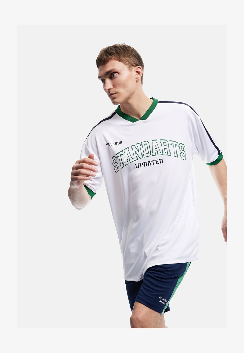 Junger Mann in weißer und grüner Sportbekleidung läuft, trägt ein Shirt mit der Aufschrift „STANDARTS UPDATED“ und marineblaue Shorts mit grünem Streifen.