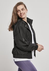 Svart lett jakke med glidelås foran, elastiske mansjetter og justerbar hette. Båret over en hvit skjorte og lilla leggings.