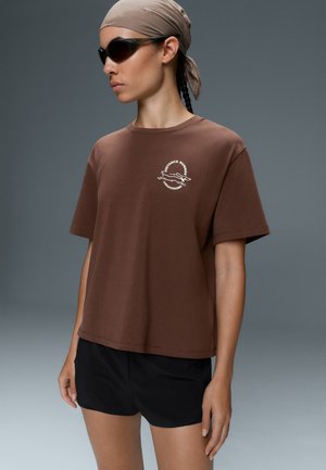 Femme portant un t-shirt marron avec le logo "Distance Runner", un short noir, un foulard beige et des lunettes de soleil noires, debout sur fond gris.