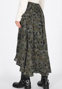 Donna che indossa una lunga gonna floreale verde e nera, un maglione bianco a maglia e stivali di pelle neri con fibbie argentate, in piedi davanti a uno sfondo bianco.