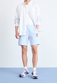 Person som bär ljusblå Nike-shorts, vit tanktopp, vita och blå sneakers och en oformad vit skjorta med blå ränder som står på ett blått golv.