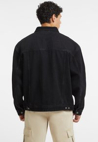 Giubbotto di denim nero con vestibilità rilassata, cuciture visibili e dettagli a bottoni sui polsini. Presenta un colletto classico e un design con yoke sul retro.