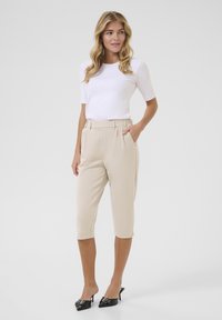 Beige capri pants yang disesuaikan dengan pinggang tinggi dan kantong samping, dipasangkan dengan atasan putih lengan pendek yang pas dan sepatu mule hitam bertumit.
