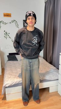 Mladý človek so čiernou čiapkou a mikinou s bielou grafikou, voľné vyblednuté jeansy, stojí v spálni s posteľou a sivými závesmi v pozadí.