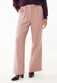 Donna che indossa pantaloni palazzo rosa cipria, stivali con tacco scuri e un maglione di lana viola scuro, con una mano in tasca.