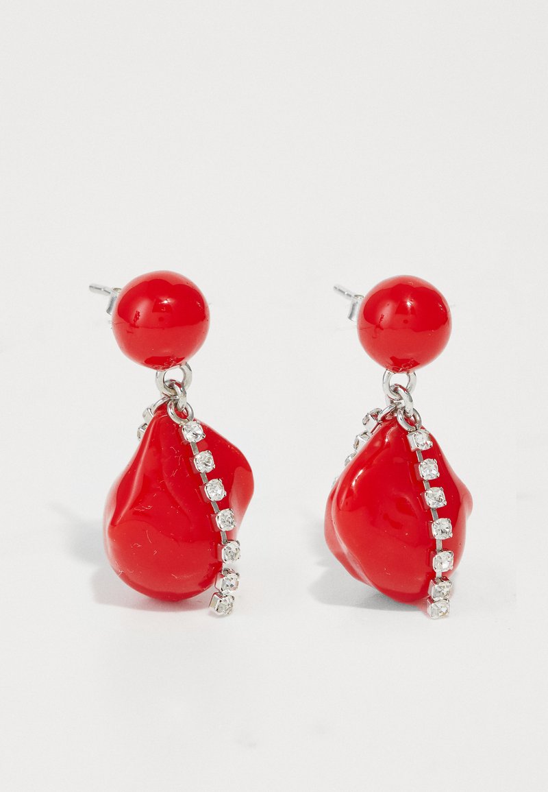 Κόκκινα κρεμαστά σκουλαρίκια με γυαλιστερές σταγόνες, στρογγυλή κορυφή και μια σειρά καθαρών πολύτιμων λίθων στο πλάι. Περιλαμβάνονται ασημένιες λεπτομέρειες.