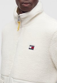 Άνδρας που φοράει λευκό fleece μπουφάν με κίτρινο φερμουάρ και μικρό λογότυπο Tommy Jeans στο στήθος.