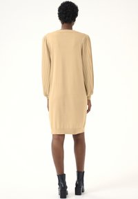 Robe pull en tricot de couleur beige doux, avec de longues manches bouffantes, une texture côtelée et un ourlet inférieur ajusté. Associée à des bottines noires.