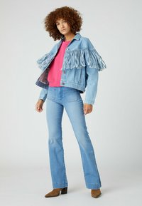 Giacca di jeans azzurro chiaro con dettagli in frange, abbinata a un felpa rosa e jeans a zampa di elefante azzurro chiaro. Include stivali con tacco marroni.