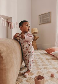 Bombažna kombinezona z rožnatimi pentljami na belem, v kombinaciji z dolgimi rokavi roza majice. V roki ima zaobljeno leseno igračo, stoji ob bež kavču.