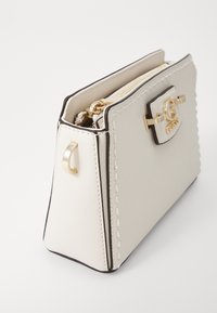Borsa strutturata beige chiaro con hardware dorato, zip superiore e superficie testurizzata. Presenta dettagli cuciti e un accento con logo.