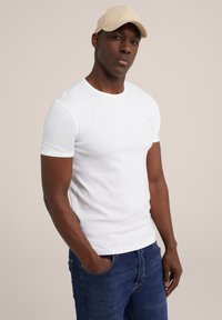 Weißes kurzärmliges Rundhalsausschnitt-T-Shirt aus Baumwolle, kombiniert mit blauen Jeans, die eine lockere Passform haben und die Hände in den Taschen stecken.