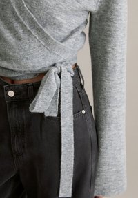 Szary sweter typu wrap z długimi rękawami, wykonany z teksturowanej tkaniny, zawiązywany paskiem. W połączeniu z czarnymi, dżinsowymi spodniami z wysokim stanem.