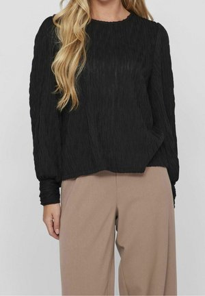 Femme portant un haut noir texturé à manches longues et un pantalon beige taille haute à coupe ample, sur un fond gris uni.
