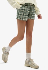 Pantalones cortos de cuadro verde y beige con cintura elástica, bolsillos laterales y un ajuste relajado, combinados con zapatillas grises y calcetines blancos.