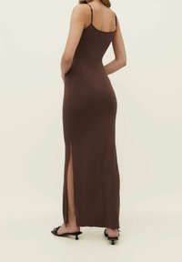 Kobieta w dopasowanej brązowej, bezrękawnej sukience maxi z bocznym rozcięciem oraz czarnych sandałach na kocim obcasie, stojąca na jasnym tle.