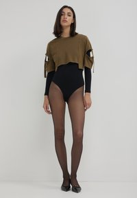 Donna con un body a maniche lunghe nero e collant trasparenti, un top verde oliva corto con maniche aperte e ballerine nere a punta, in piedi contro un muro bianco.