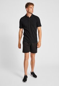 Schwarzes Polo-Shirt, kurzärmlig, aus glattem Stoff. Kombiniert mit schwarzen Shorts mit roten Akzenten. Getragen mit schwarzen Sportschuhen.