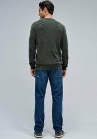 Man standing facing away, wearing a dark green sweater, blue jeans, and gray sneakers against a plain gray background.