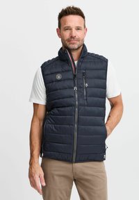 Navy blue, puffy, sleeveless vest with a zip front, zippered chest pocket, and vertical quilting. Features a logo patch on the left chest.