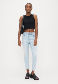 Γυναίκα που στέκεται μπροστά σε λευκό φόντο, φοράει μαύρο αμάνικο crop top, ανοιχτό μπλε skinny τζιν, μαύρα loafers και κρατά μαύρη τσάντα ώμου.