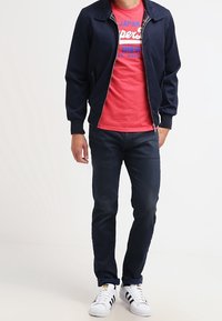 Giacca bomber navy, t-shirt grafica rossa con scritte blu, jeans blu scuro e sneakers bianche con strisce nere e dettagli gialli.