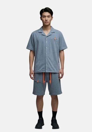 Homme portant une chemise bleue à manches courtes assortie avec boutons et un short avec cordons orange, chaussettes noires et baskets noires, debout droit.