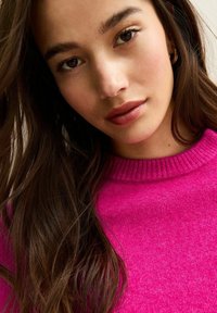 Jasnoróżowy sweter z prążkowanym dekoltem, charakteryzujący się miękką fakturą. Długie, ciemne włosy częściowo oprawiają twarz, podkreślając naturalne rysy.