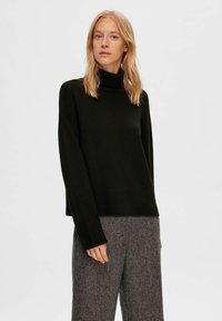 Un pull à col roulé vert foncé en tissu tricoté doux avec des poignets et un ourlet côtelés. Assorti à un pantalon gris à motif pied-de-poule.
