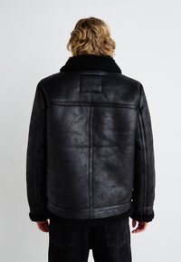 Black faux leather jacket with a shearling collar and cuffs, featuring a smooth texture and a square patch on the upper back.