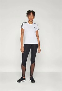 Camiseta deportiva blanca con franjas negras en los hombros, combinada con leggings negros que tienen detalles de malla y franjas blancas a los lados. Zapatillas negras.