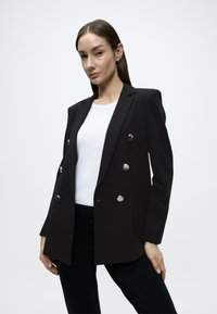 Μαύρο σακάκι με διπλή σειρά κουμπιών σε ασημί απόχρωση, με αιχμηλές πέτο και δομημένη σιλουέτα, φορεμένο πάνω από λευκό πουκάμισο και μαύρο παντελόνι.