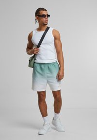 Fehér ujjatlan felső, zöld és fehér színátmenetes sort, pálma mintával, fehér sportcipő, kicsi zöld crossbody táska, szürke háttér.