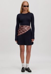 Marineblaues, geripptes Strickkleid mit langen Ärmeln, das einen diagonalen Karo-Stoffakzent an der Taille aufweist, kombiniert mit schwarzen Slippern und weißen Socken.