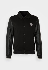 Sort jakke med stofkrop og læderærmer. Har knaplukning foran, stribede ribbenmanchetter og et broderet logo på brystet.
